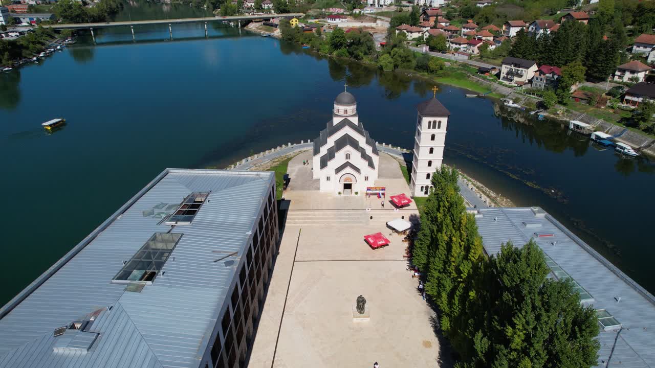 Drone perspective of the Church of Saint Tsar Lazarus in Andricgrad, Visegrad, Bosnia and Herzegovina, with riverside setting, bell tower, nearby modern structures, and scenic residential surroundings