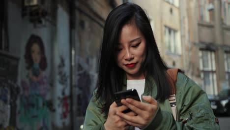 Attraktive-Junge-Frau-In-Stilvollem-Outfit,-Die-Mit-Interesse-Auf-Dem-Smartphone-Chattet-Und-Tippt-Und-Mit-Graffity-Auf-Der-Straße-Lächelt