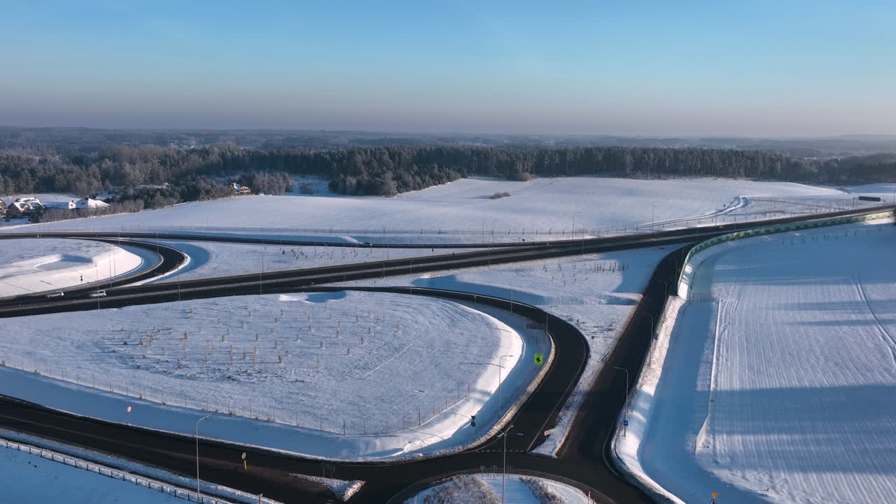 Luftoptagelser af et motorvejskryds omgivet af snedækkede marker og skove under en klar, blå himmel. Jævn panorering med kameraet.