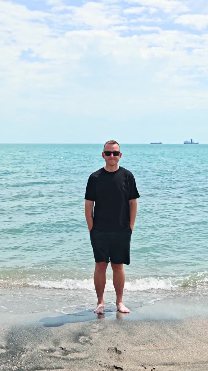 Uomo in piedi sulla spiaggia con l'acqua intorno ai piedi, mentre si vedono barche sullo sfondo in una giornata limpida