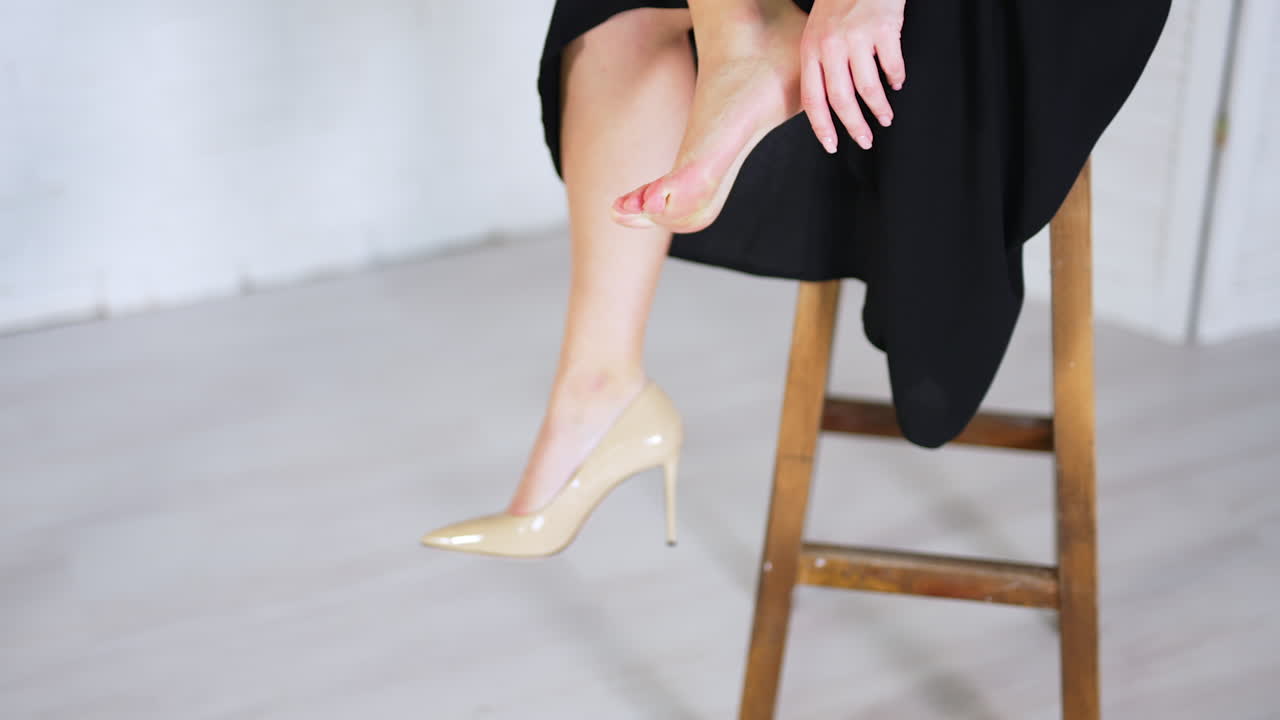 Unknown woman sits on a high tabouret and takes off her shoe. Lady in long dress massages her foot tired from high heels. White blurred backdrop.
