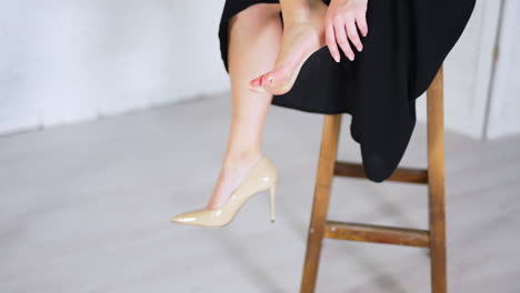 unknown woman sits on a high tabouret and takes off her shoe. lady in long dress massages her foot tired from high heels. white blurred backdrop.