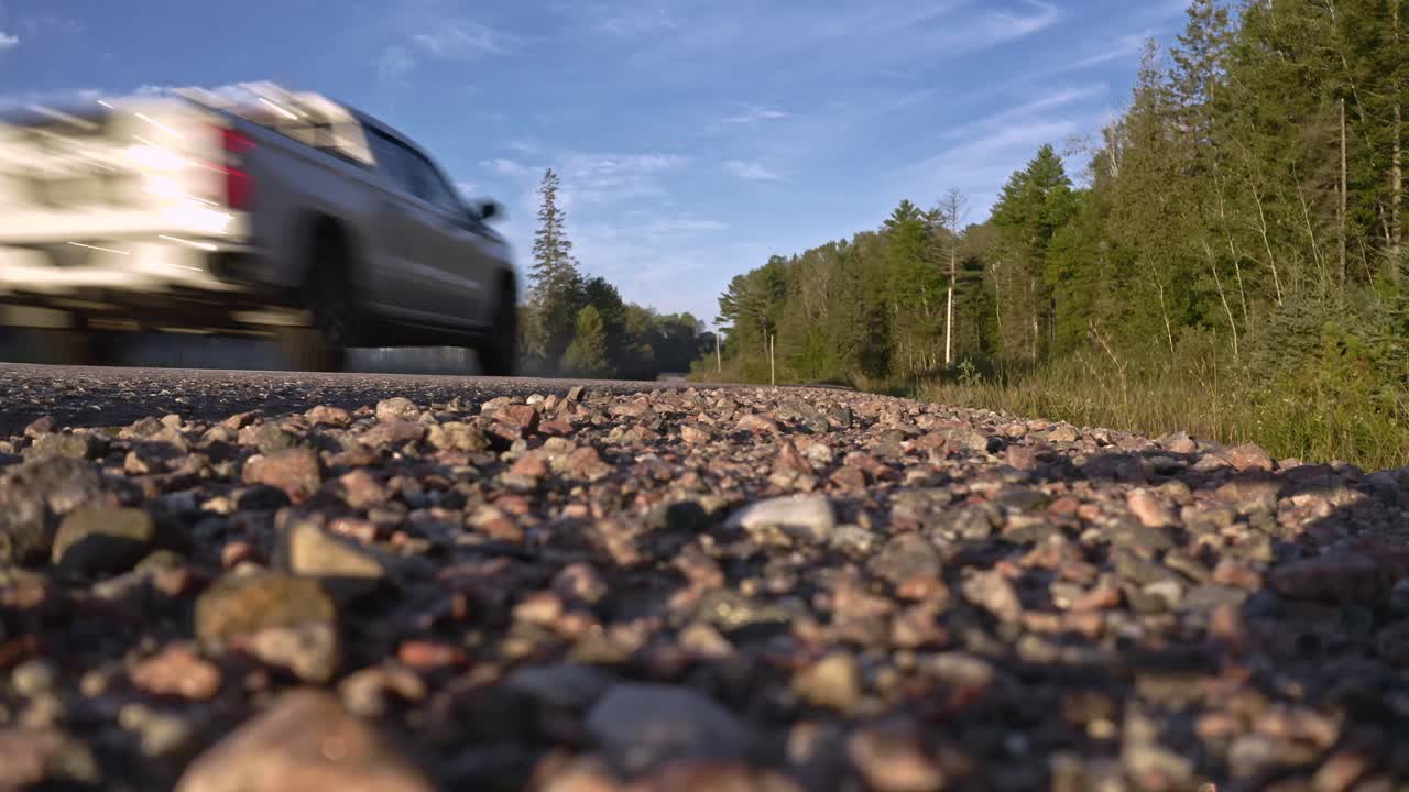 Ground level shot of truck driving by on rural highway with gravel shoulder in foreground and forest in the background