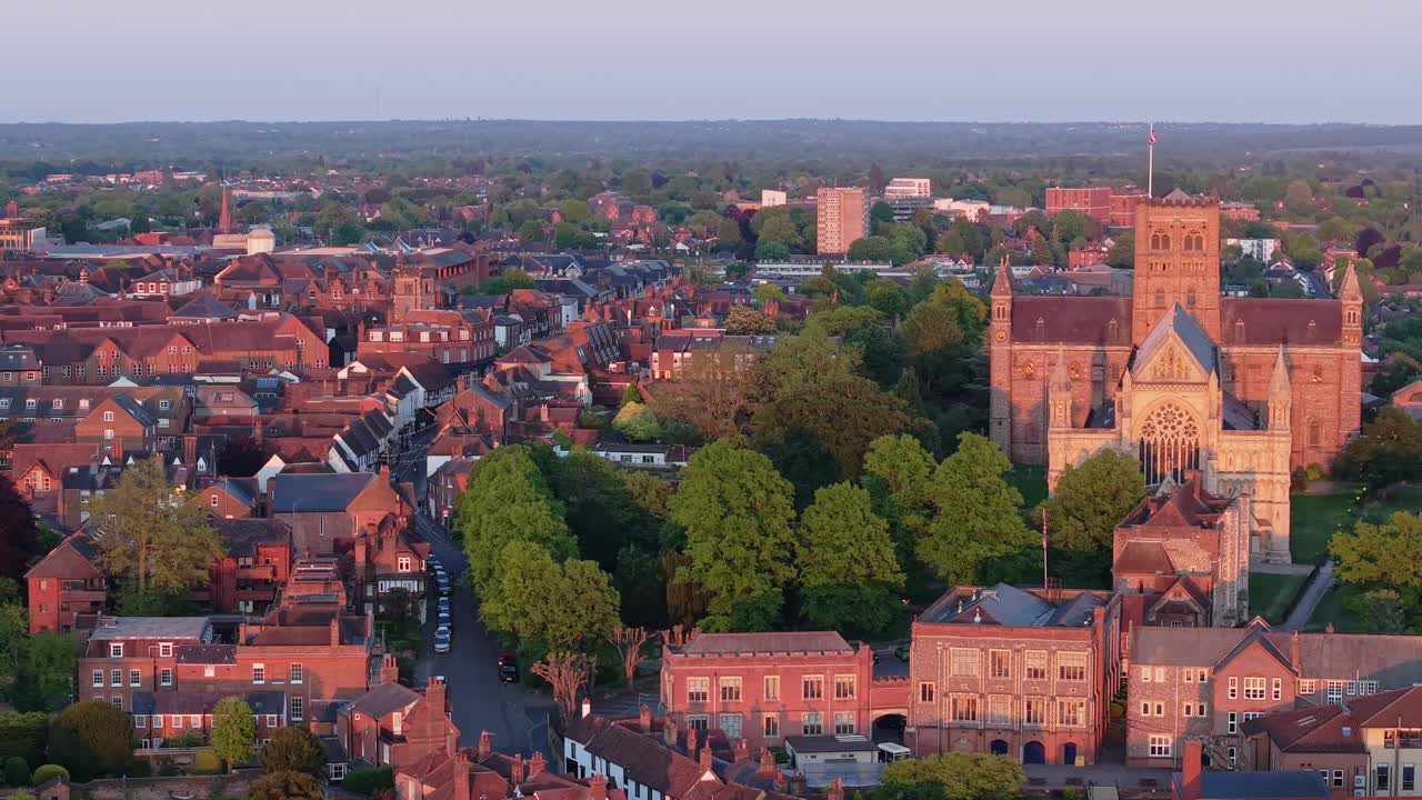 A cinematic parallax drone shot sweeps over St Albans city, gradually revealing the majestic cathedral rising above the urban landscape