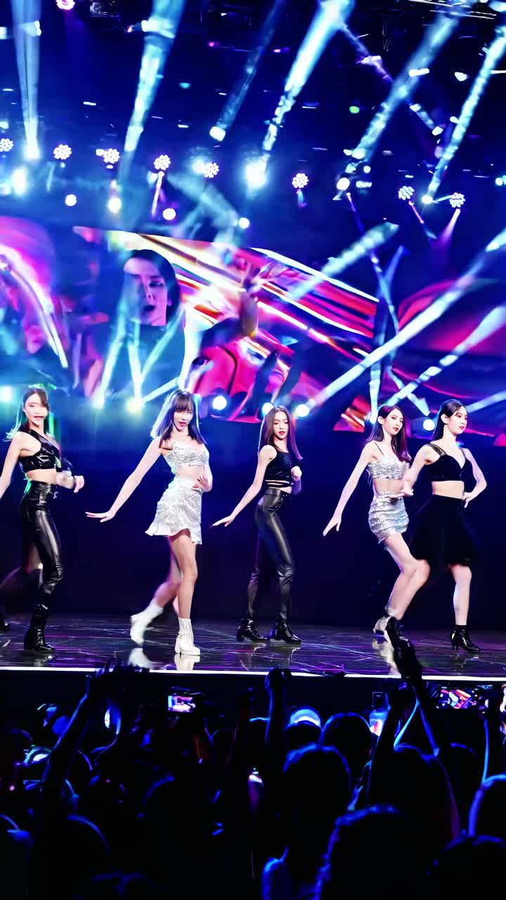 Group of young female kpop idols dancing and singing on stage during a live concert in front of a large led screen, with fans cheering and holding smartphones recording the show