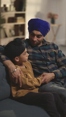 vertical video of loving sikh father and young son wearing turbans sitting on sofa at home talking together