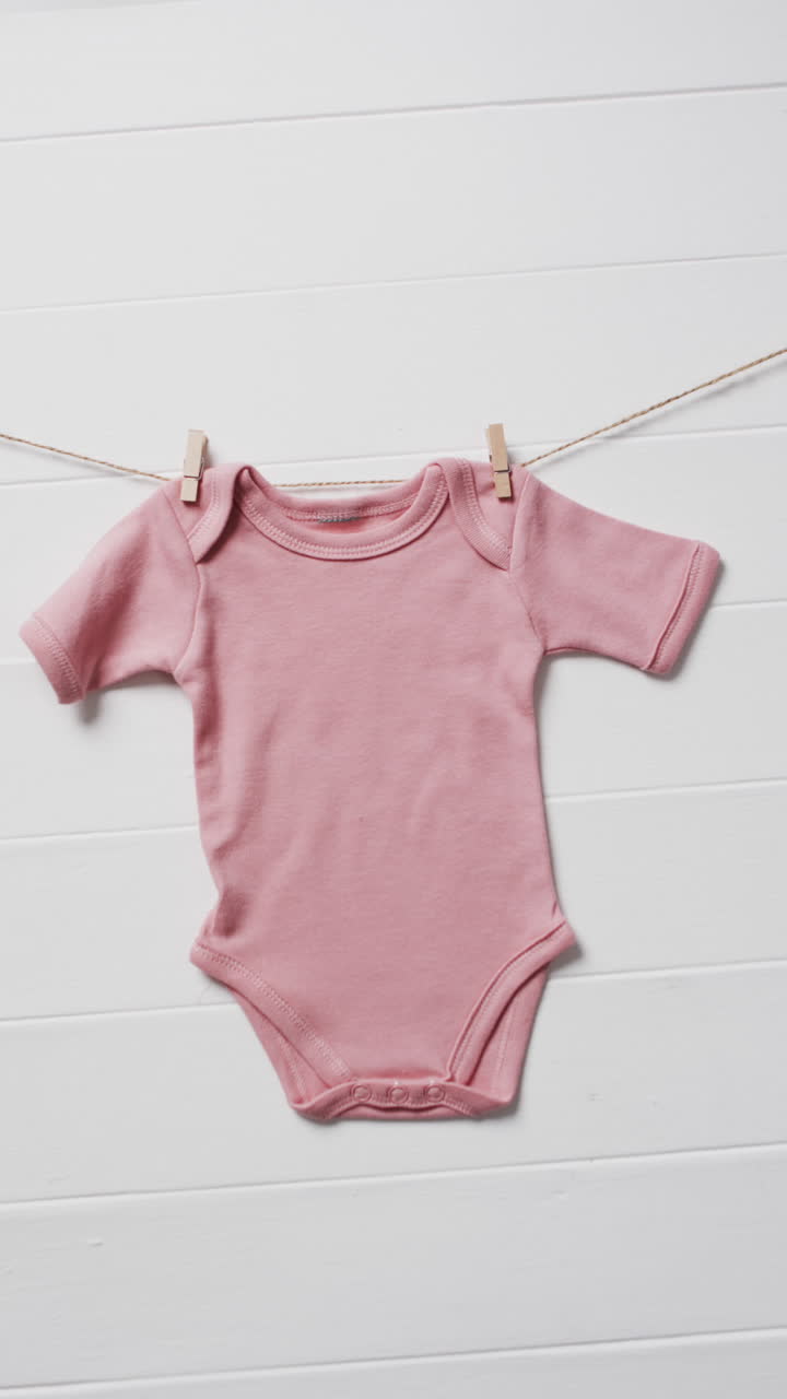 Vertical video of pink baby grow hanging on clothes pegs with copy space on white background