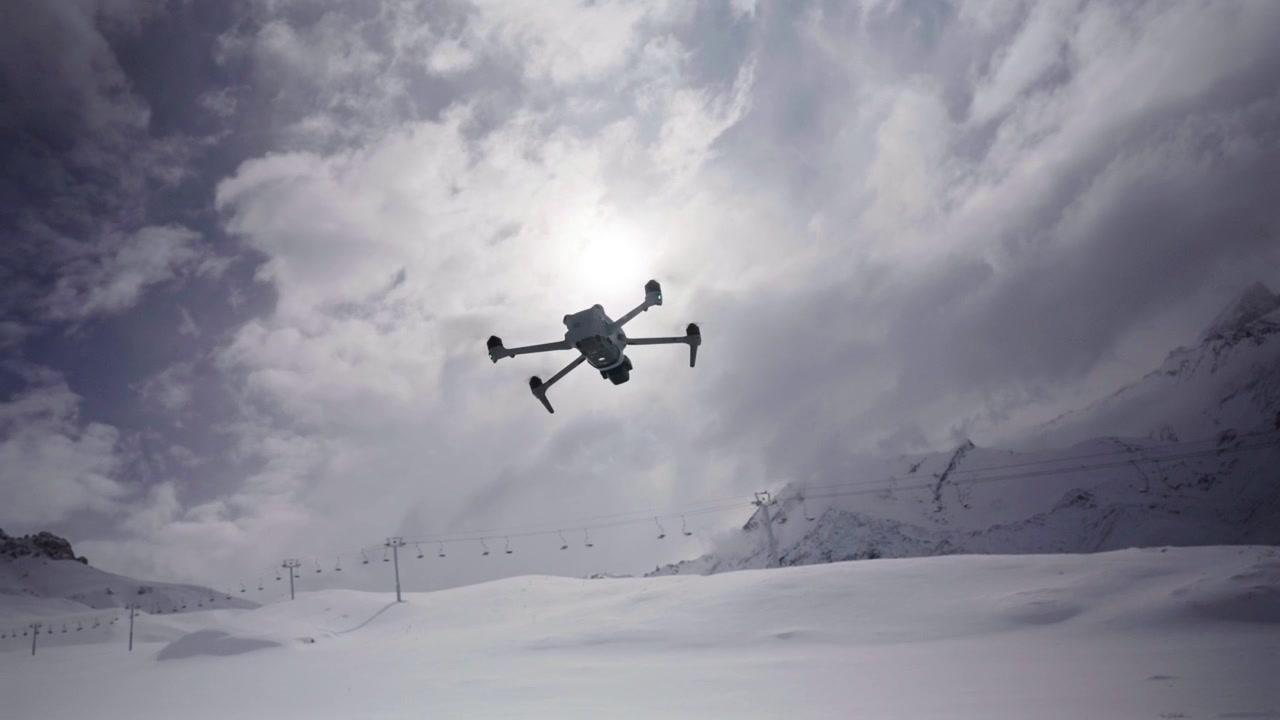 Quadcopter drone hovers above a snow covered mountain landscape with ski lift towers, alpine terrain and dramatic winter clouds, showcasing aerial technology, slow motion orbit.