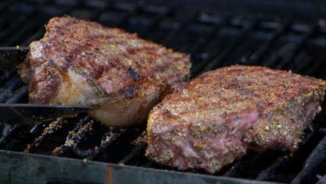 A-pair-of-meat-tongs-lift-a-nearly-cooked-juicy-rib-eye-steak-on-a-grill-and-presses-on-it-and-another-steak