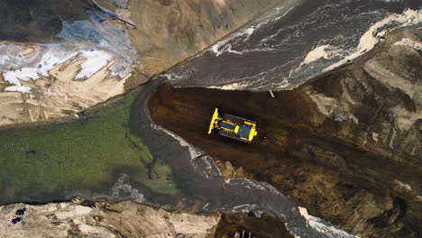 top-down aerial shot of yellow bulldozer operating and moving soil in front of river channel at worksite