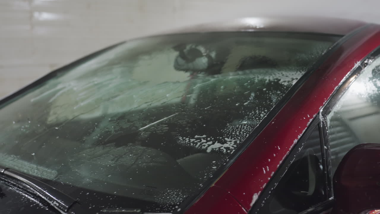 Rear view of person using high pressure car wash hose to rinse soap from car windshield inside garage with water droplets and suds streaming down glass surface during cleaning process