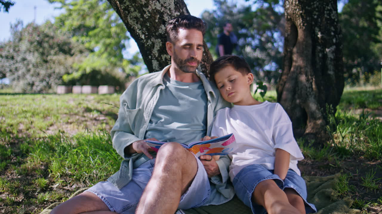Dad kid watching book sitting green grass at forest. Loving parent son reading