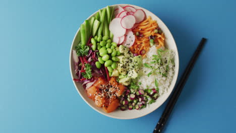 Composition-of-bowl-of-rice,-salmon-and-vegetables-with-chopsticks-on-blue-background