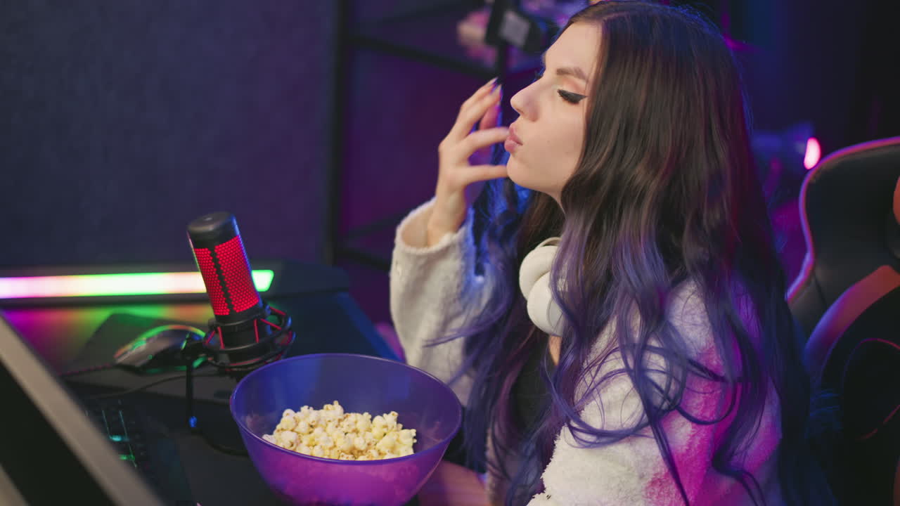 Young girl seated in vibrant home studio finishes eating popcorn, wipes fingers before resuming work at microphone and screen, ambient colored lights