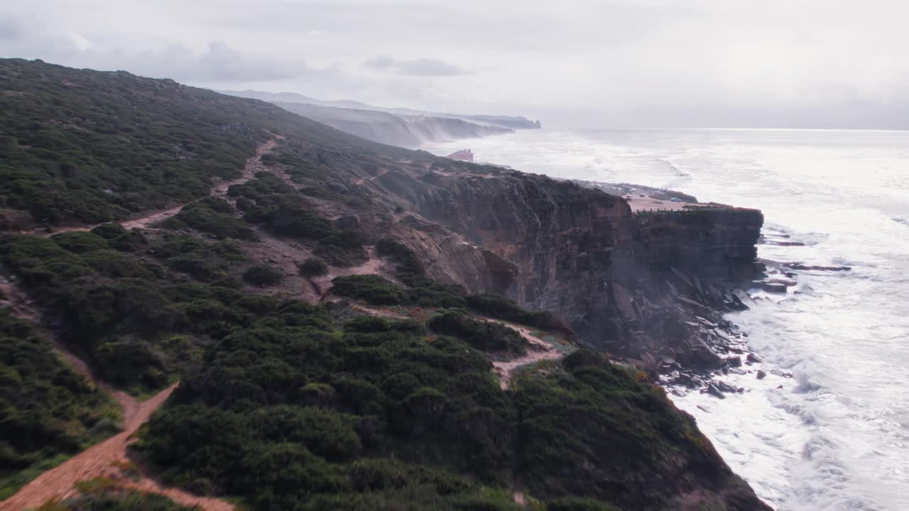 Misty aerial view of dramatic cliffs and winding coastal path near Cabo da Roca, Portugal. Crashing Atlantic waves and rugged terrain evoke wild beauty at Europe’s westernmost point