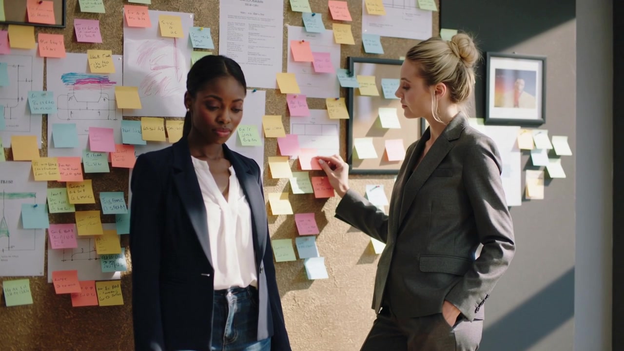 Reaching during review, right woman grey-suited adjusting pink note on corkboard colleague watching