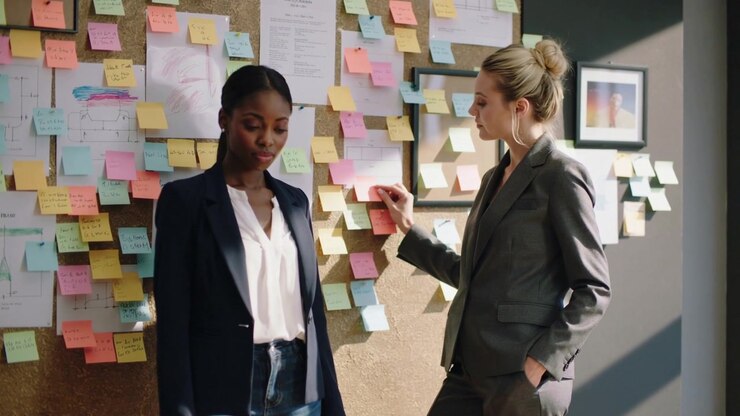 Reaching during review, right woman grey-suited adjusting pink note on corkboard colleague watching