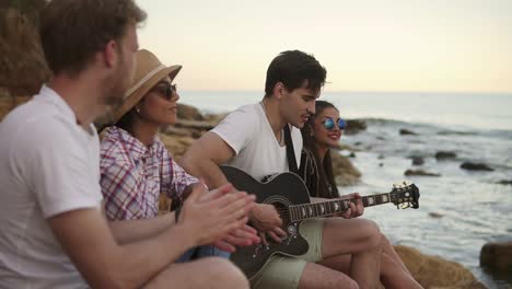 group of young hipster friends sitting on the rocks by the seashore and playing guitar and singing songs. slow motion shot