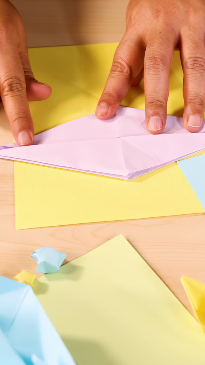 Mani piegano con cura carta origami color pastello su un tavolo di legno sotto un'illuminazione brillante e uniforme.