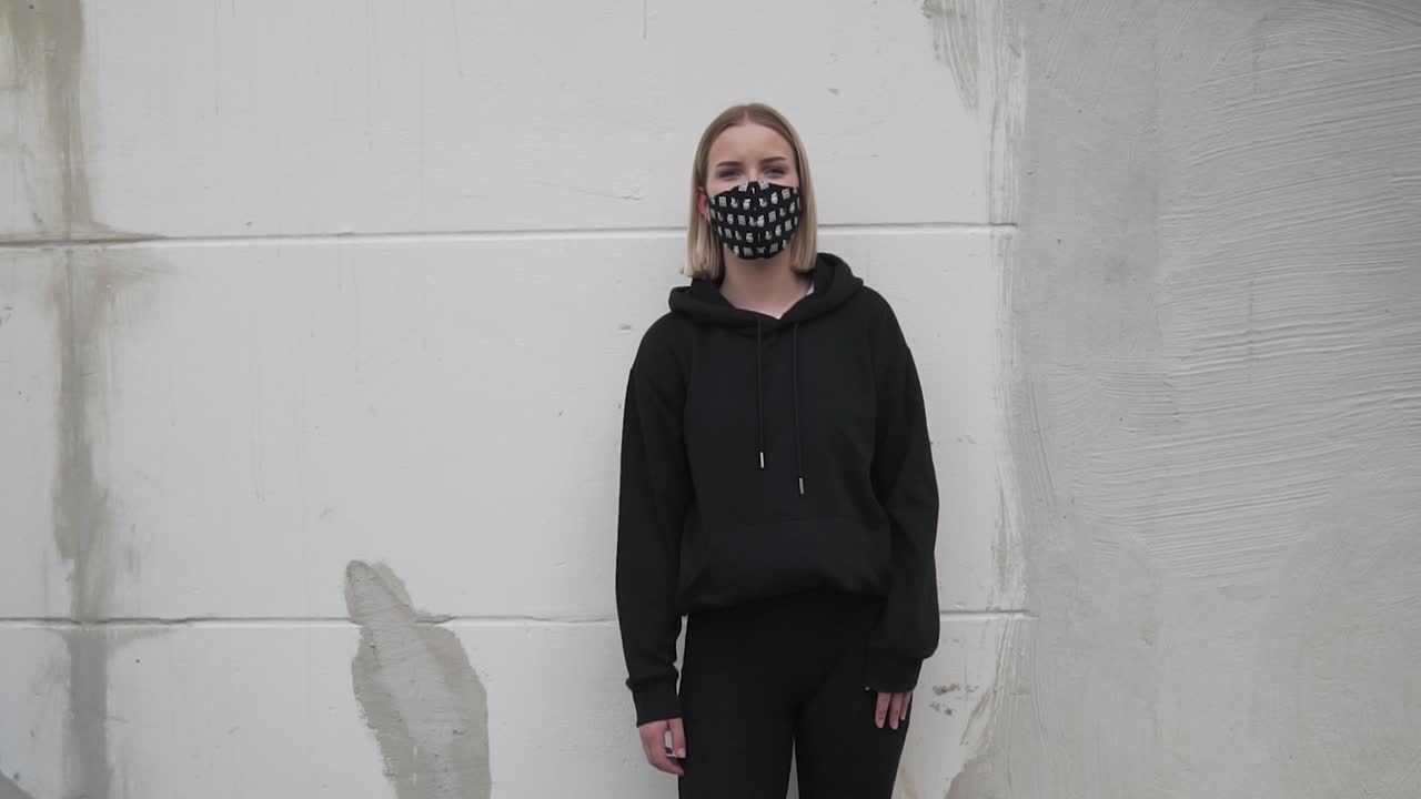 молодая женщина в черной одежде и маске на лице стоит у стены и смотрит в камеру