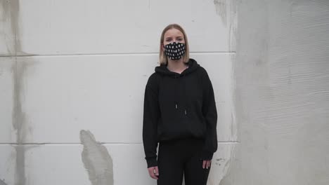 young female in black clothes and face mask standing by wall looking at camera