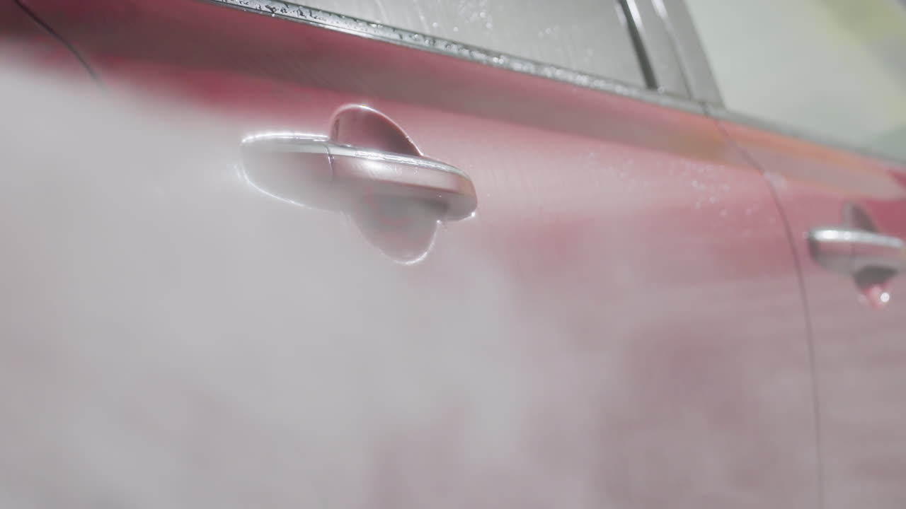 close up of red car door being rinsed by high pressure water spray inside garage wash bay revealing glossy metal surface covered in suds and droplets with dynamic flow and shining reflection