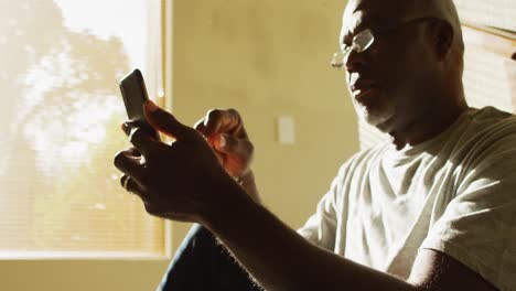 Thoughtful-african-american-senior-man-sitting-at-home-in-the-sun-using-smartphone