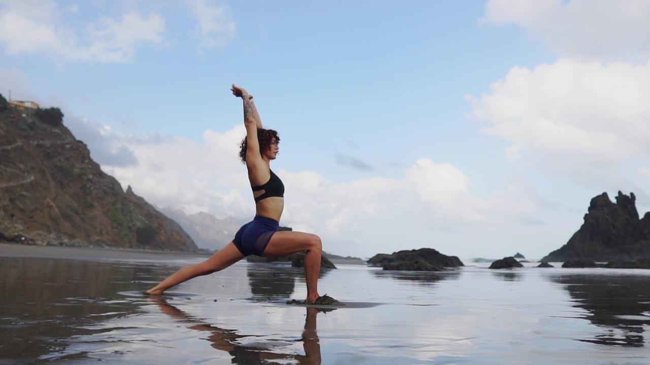 Slow motion footage shows a young, slim woman practicing stretching and yoga, standing near the ocean and looking out into the distance