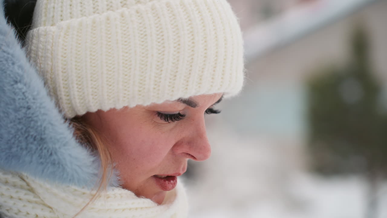 La mamma sulla neve trattiene il respiro, in attesa. Primo piano di una madre con cappello di lana color crema, sciarpa e cappotto blu con cappuccio, mani vicino al viso, occhi fissi in lontananza, un accenno di lacrima sulla guancia, espressione resiliente.