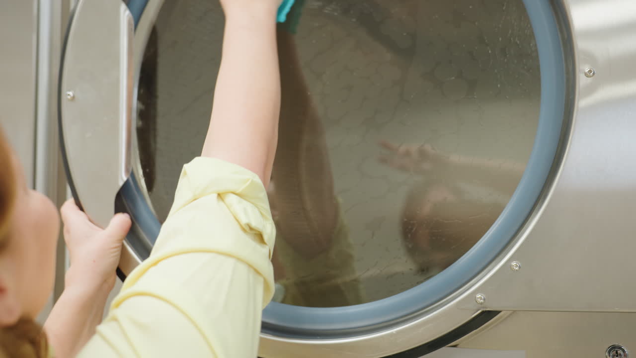Close up of laundry worker thoroughly cleaning transparent surface door of washing machine with soapy towel, focusing on removing dirt and maintaining hygiene inside laundromat facility