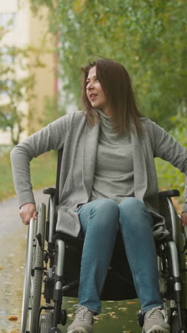 young woman with paralysis enjoys rest in green park on nasty day. pretty lady in wheelchair smiles contentedly looking into distance. recovery from injury