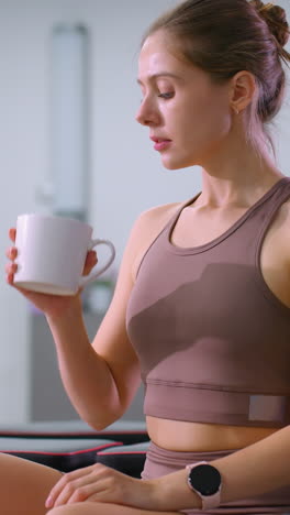 la señora se toma un descanso del ejercicio, bebiendo café mientras está sentada en el gimnasio, un gran espejo detrás refleja su postura relajada, el equipo de gimnasio y las esteras de fitness añaden a la atmósfera de bienestar