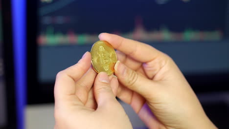 unrecognizable woman holding and looking at bitcoin in her fingers in front of cryptocurrency trading graphs