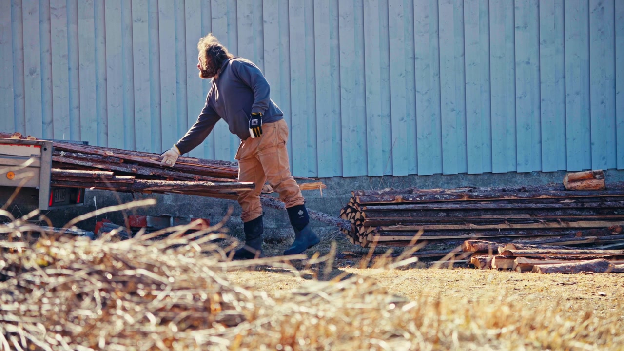 Man Pulls A Long Wooden Plank From A Stacked Pile Of Lumber Outdoors, Showing Manual Work. Static Shot