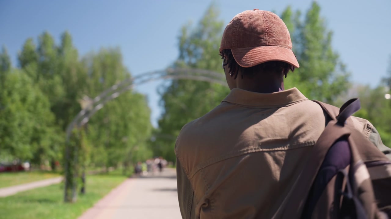 Close up of man walking away from camera through outdoor park on sunny day wearing casual clothing and carrying backpack. scene includes green trees distant buildings parked cars along path