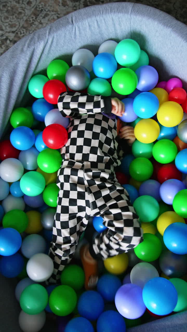 Funny kid in checkered pajama hiding in the balls of dry pool. Happy energetic toddler tumbles in the basin and gets out of it. Top view.