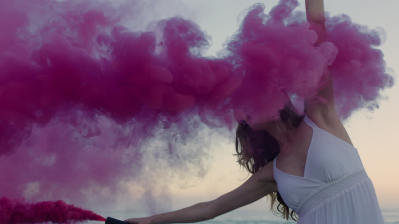 bellissima donna che agita una granata di fumo rosa che balla sulla spiaggia al mattino presto celebrando la libertà creativa