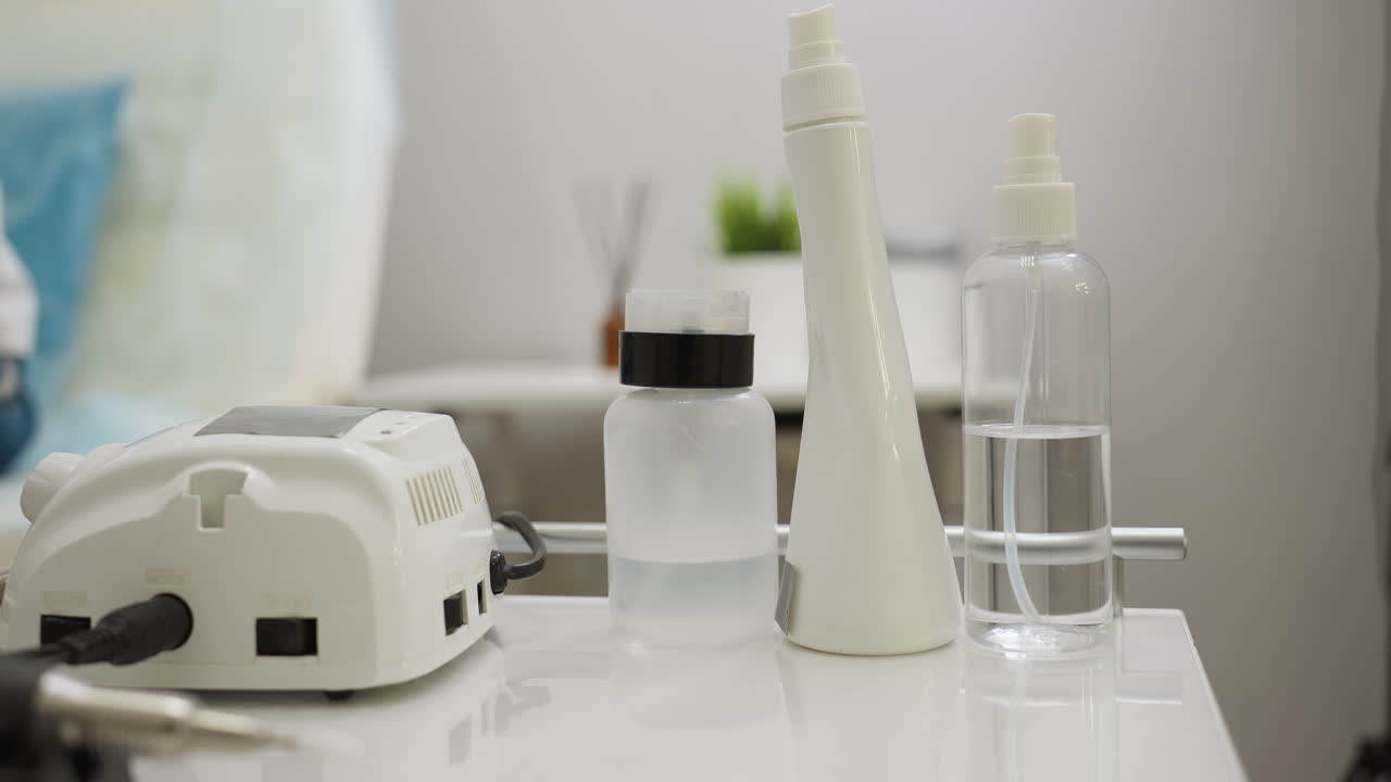 Close-up view of essential tools and bottles arranged neatly on white table, including electric device, spray bottles, and containers, with blurred background showing green indoor plant