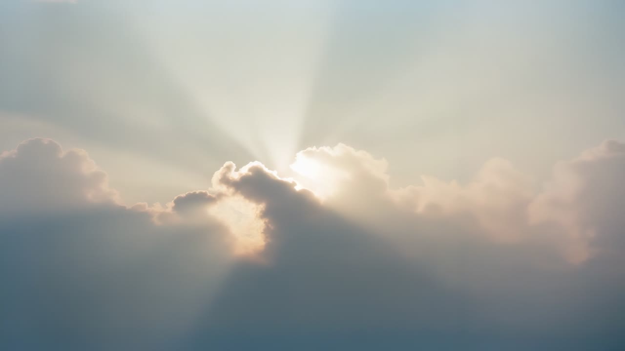 Sun emerging behind central cumulus cloud bank, casting crepuscular rays across pastel sky at dawn