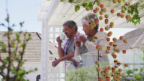 animation of emoji icons over senior biracial couple drinking coffee on balcony