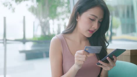 beautiful asian young female with cheerful expression, holds smart phone and credit card, banks online,happy young girl holding plastic credit card while using mobile phone at the cafe