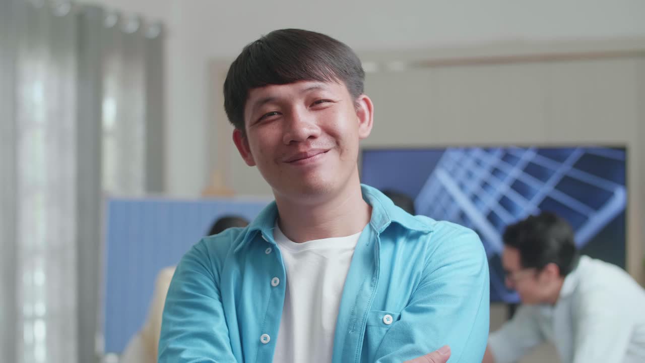 미소 짓는 아시아 남성 엔지니어가 동료들이 사무실에서 태양전지 작업에 대해 논의하는 동안 카메라에 팔을 고 있습니다.