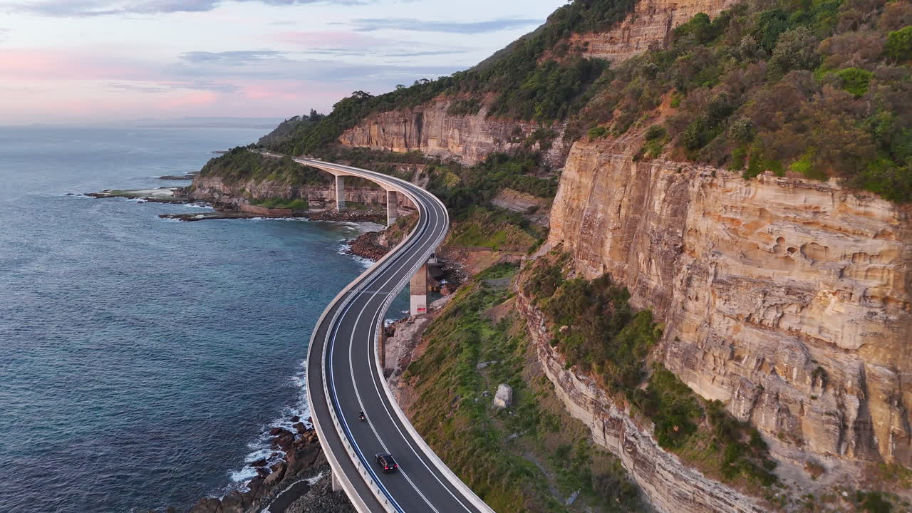 Drone footage of a car moving along the winding Sea Cliff Bridge, surrounded by dramatic rock formations and the Pacific Ocean. Cinematic aerial footage.