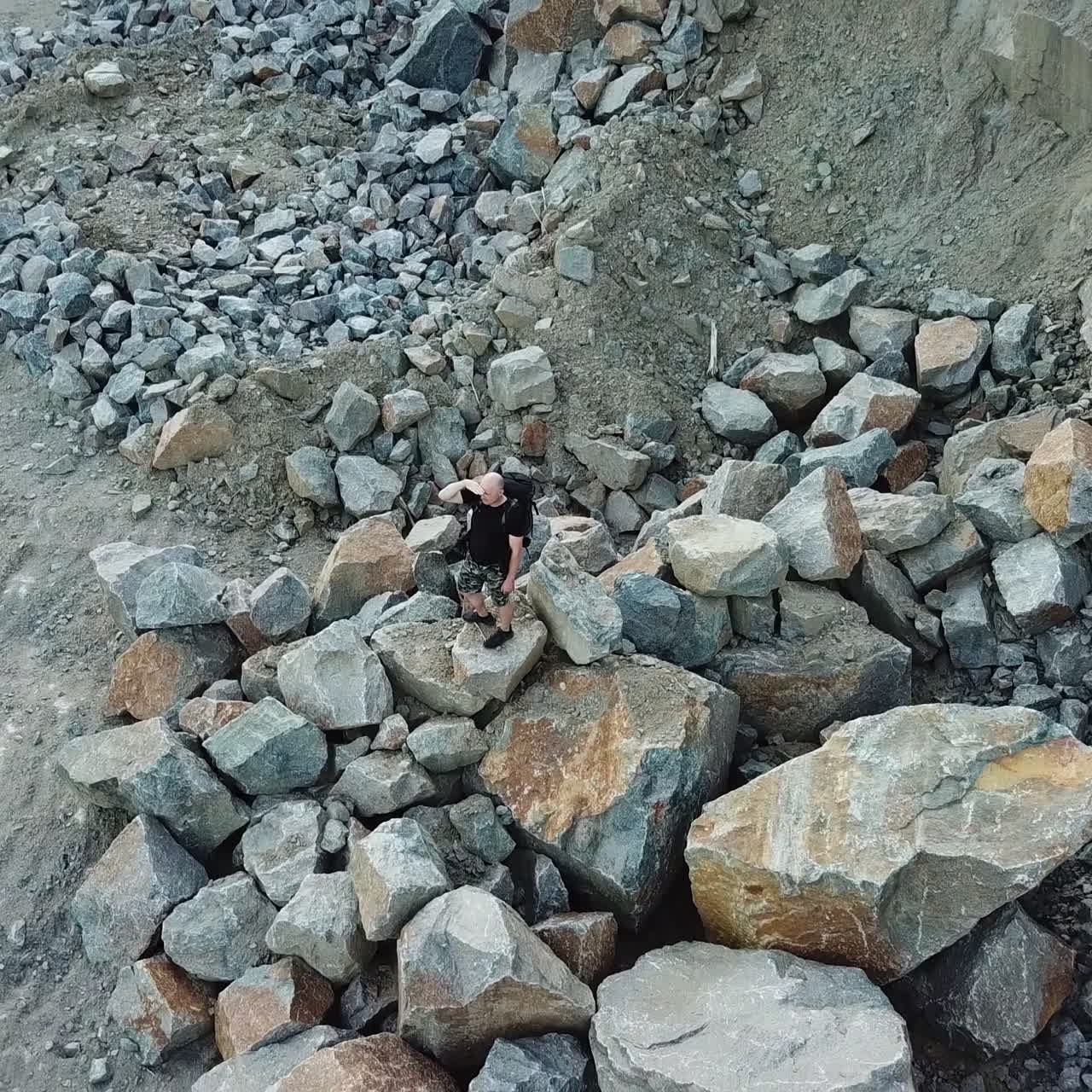 A tourist in shorts and a backpack is standing on a huge stone in the middle of a quarry and looking into the distance to the surrounding landscape. Camera motion up. Aerial view.