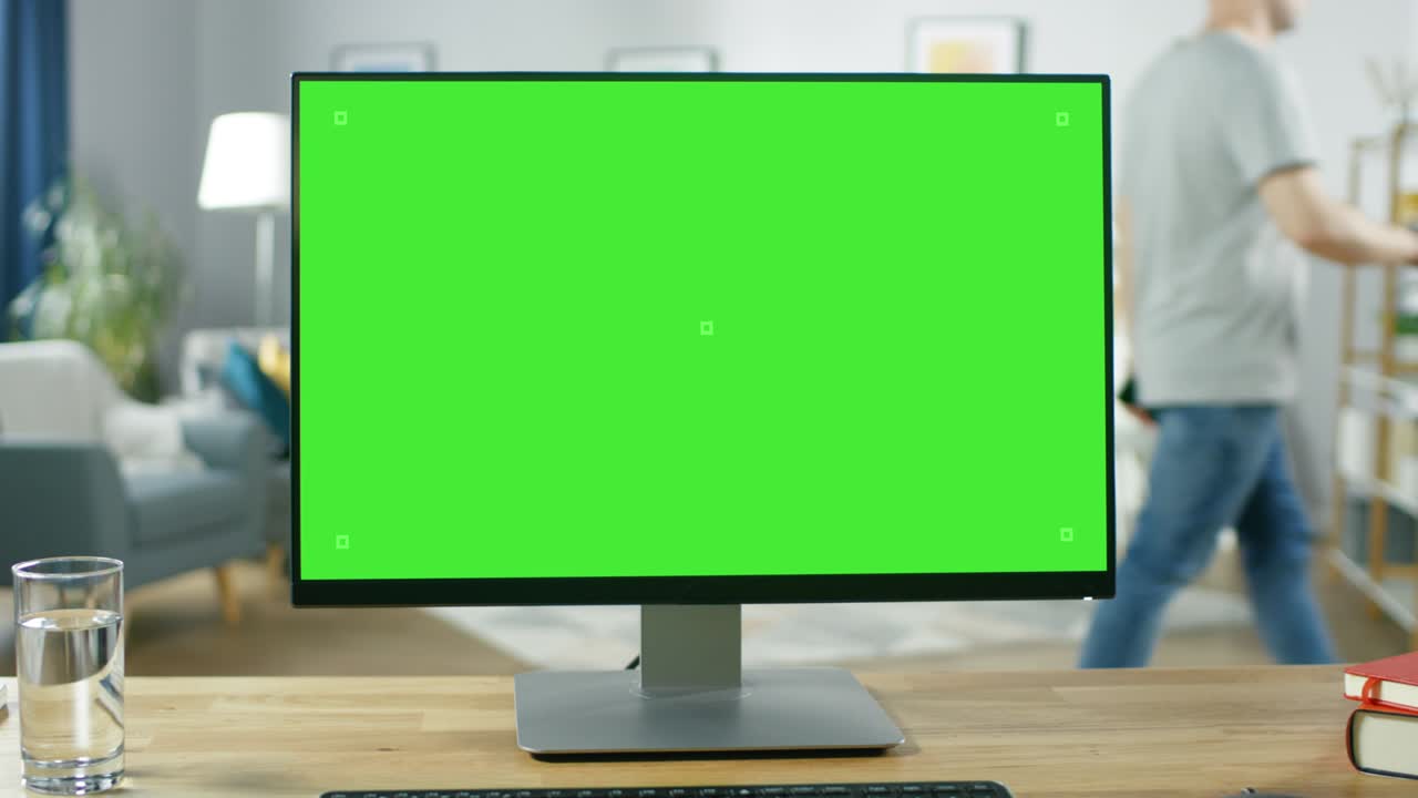 초록색 모형 화면 디스플레이를 가진 현대적인 개인 컴퓨터가 아한 거실의 책상 위에 서 있습니다. 드폰을 들고 있는 남자가 그의 아파트를 고 있습니다.
