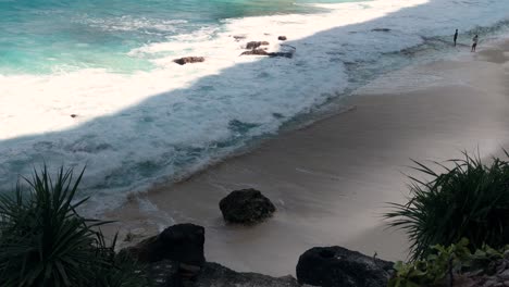 Luftaufnahme-Des-Kristallblauen-Wassers-Des-Diamond-Beach-In-Nusa-Penida,-Indonesien,-Das-Auf-Die-Küste-Trifft,-Mit-Ein-Paar-Touristen,-Die-In-Der-Ferne-In-Den-Wellen-Stehen