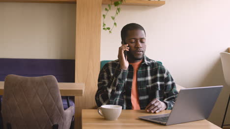 hombre sonriente hablando por celular mientras trabaja con una computadora portátil sentada en la mesa del café