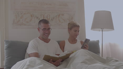 man reading book in bed, woman looking at phone screen at home