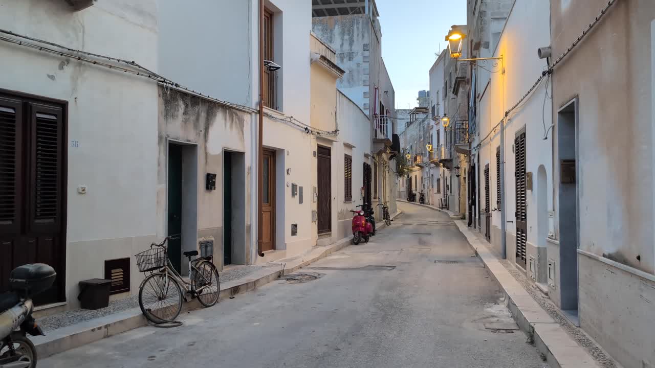 Slow pan right along a narrow street in Favignana, Sicily at dusk. Empty road with bikes, a Vespa, and warm lights creating a calm Mediterranean evening scene