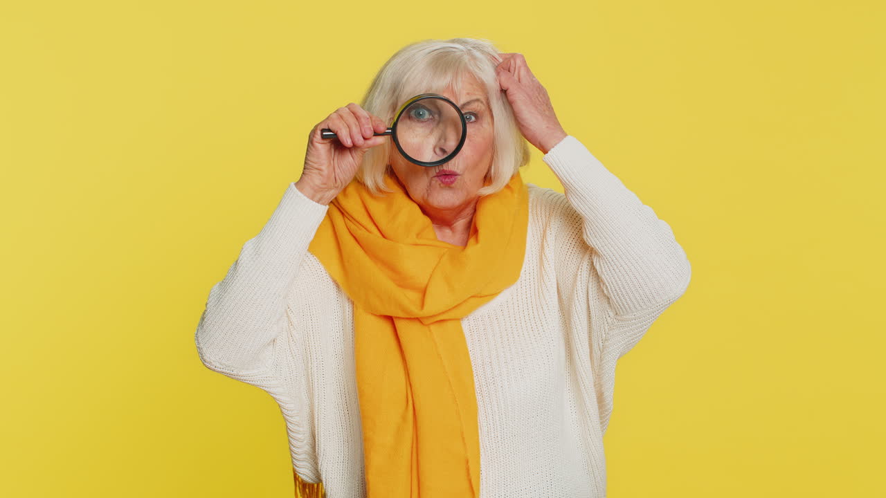 Senior old woman holds magnifying glass near face looking at camera with big zoomed eye analyzing
